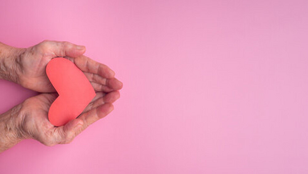 People, age, love, and health care concept. Red paper cut a heart shape on the palm of a senior woman with a pink background. Healthcare, wellbeing, World heart day, World health day.